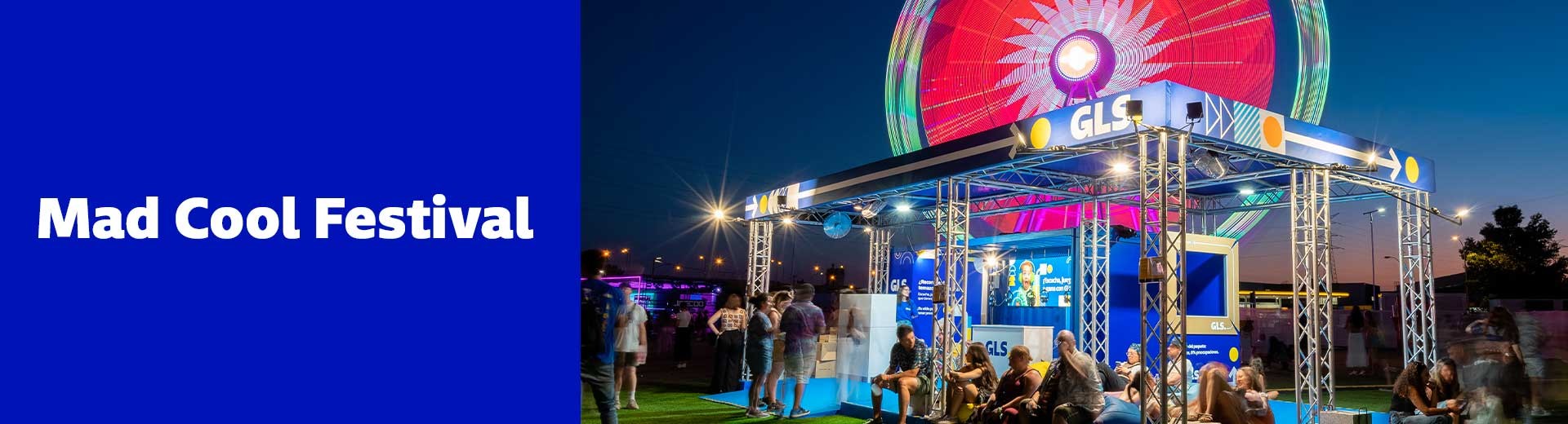Stand nocturno de GLS iluminado en Mad Cool Festival, con asistentes descansando y noria iluminada de fondo