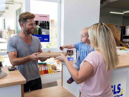 A woman with a baby collects a parcel from a shopkeeper at a GLS Pickup Point.